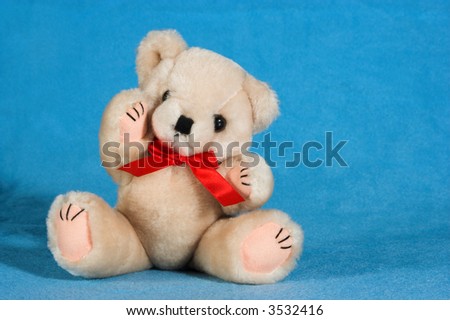 Natural colored teddy bear on a blue blanket. He has a red ribbon around his neck.  He is sitting on the blanket and has one arm raised