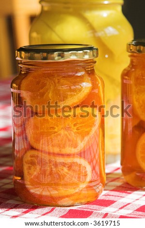 Preserved fruit in a glass jar. There are two jars of oranges and one jar of lemons in the background.  The jars are on a red and white tablecloth.