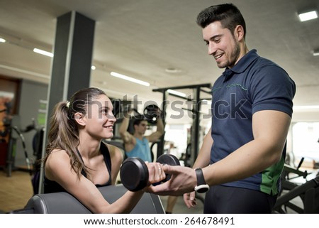 Personal trainer helps girl in gym. Man training in background.