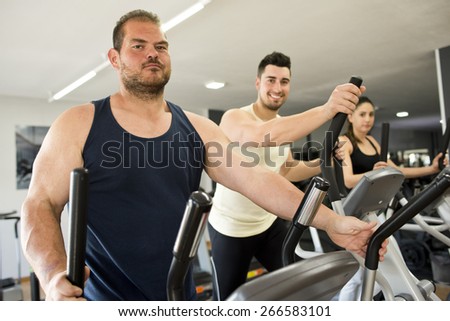 Big guy training in elliptical bike with friends at gym. Real people working body with ambient light