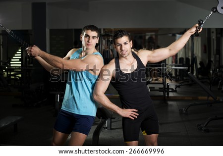 Two strong young brunette men posing at gym in pull machines