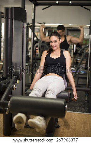 Young woman training legs at gym pull machine, man training in the background
