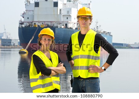 Two dockers, a man and a woman, posing in front of a huge cargo ship, moored off at an anchor buoy in an industrial harbor