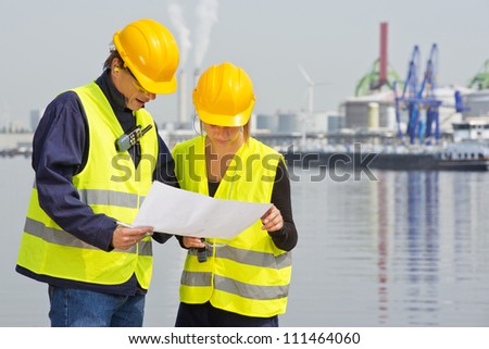 Two engineers, wearing safety gear, including goggles, hard hat, ear plugs and reflective safety vests, discussing blue prints in an industrial harbor with factories and plants in the background