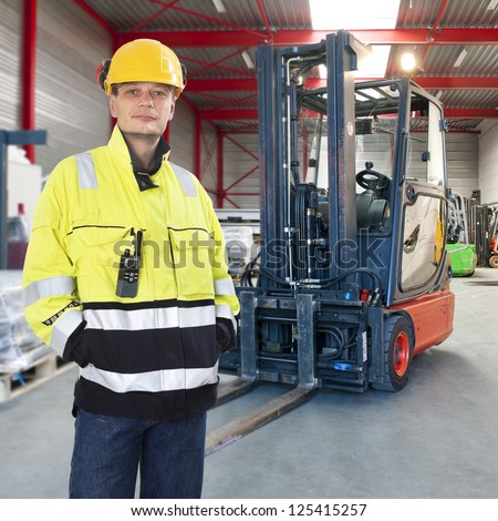 Man waring safety clothes including a hard hat standing proudly in front of his forklift truck in a warehouse Man waring safety clothes including a hard hat standing proudly in front of his forklift truck in a warehouse
