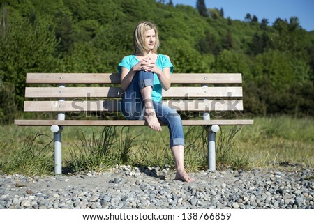 Pretty woman, sitting barefoot on a wooden bench in the park, one knee drawn up to her face, enjoying the warm spring sunlight
