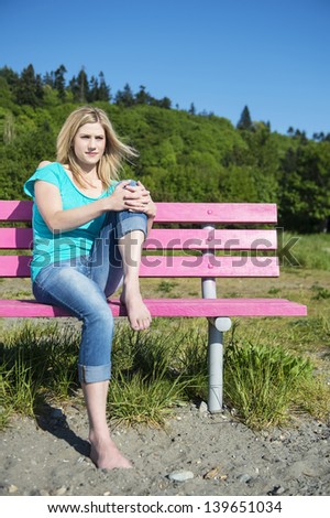 Young Woman Sitting on a bench in a park, with her hands over her knee, and her leg pulled up. Wind blowing through her hair