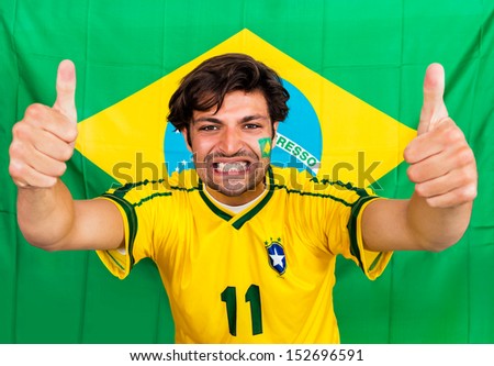 Brazilian sports fan, supporting his national team cheering with his outstreched arms and thumbs up in front of a Brazil Flag, wearing the national colors
