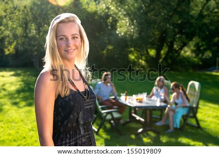 A young woman standing in a sunny garden while her friends enjoying a garden party in background