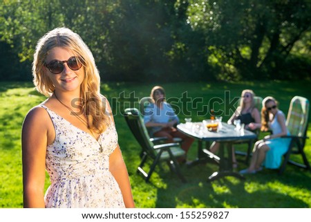 A young woman standing in a sunny garden while her friends enjoying a garden party in background
