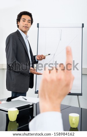 Businessman explaining graph on filpchart with colleague raising hand in conference room