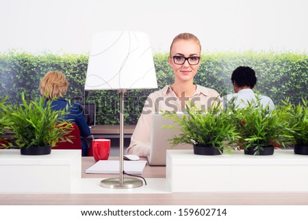 Portrait of smiling business woman working in a green office, sitting at a hot desk, with two others in adjacent seats