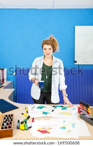 Industrial designer standing behind her desk, covered with sketches and product drawings, holding a coffee cup and smiling