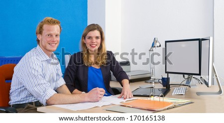 Two coworkers sitting behind a desk in front of a folder and some paperwork, with a dual monitor computer