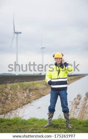 Contractor communicating in the field and calling his clients while he is standing in the grass with windmills in the background