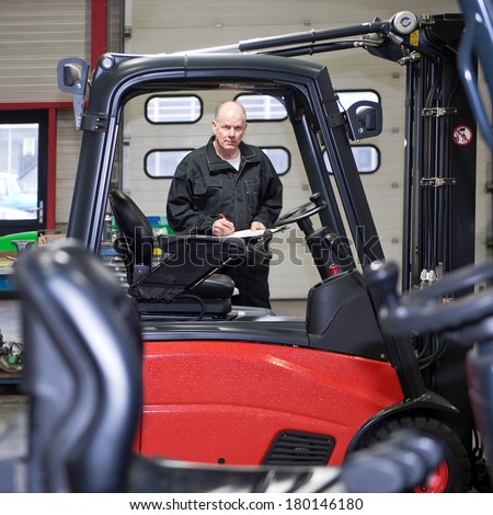 An experienced and reliable mechanic, with a clipboard and pen in his hand, working on a forklift at a garage, seen from the cabin of an adjacent forklift.