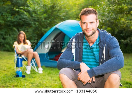Man on the camping in front of his tent, where his girlfriend sits and reads a book. enjoying vacation in the forest