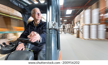 Elderly man driving a forklift through a warehouse where cardboard boxes are stored.