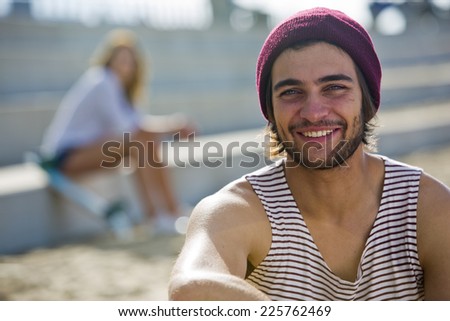 Cool dude smiling at the camera, with his girlfriend in the background