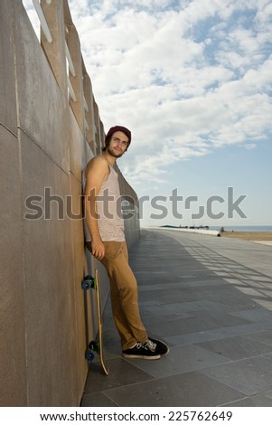 Young skater boy leaning against a granite wall of an urban boardwalk urban setting along the seashore with a skateboard next to him