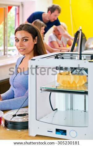 Young woman, standing next to a 3D printer at a rapid prototyping design firm, with several people working in the background