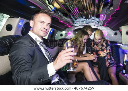 Smiling handsome young man in the back of a stretch limousine, toasting a glass of champagne towards the camera, with three people in the background