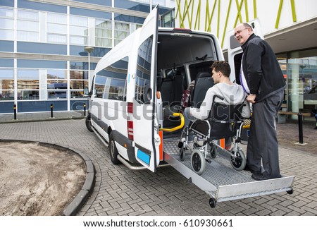 The driver of a wheel chair taxi, helping a disabled man in a wheel chair, using the lift in the back of his mini van