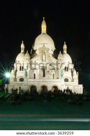 The staircase of the Sacre  Coeur in Montmartre, filled with people watching over the Parisian skyline at night, and enjoying the entertainment provided by street artists. Cars driving by