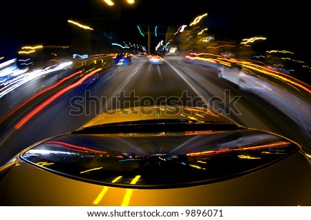A car, surrounded by blurred other cars due to the high speed, driving in a busy down town area with lots of lights, traffic and traffic lights
