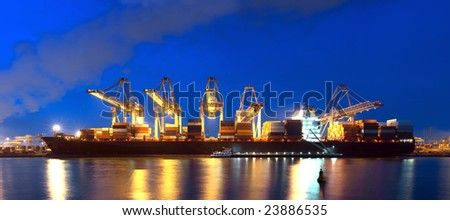 An XXXL file of a container ship being unloaded at night at a busy commercial harbour, with a small trawler moored alongside.