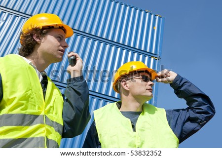 Two dock workers in safety clothing supervising and giving instructions using a walkie-talkie.