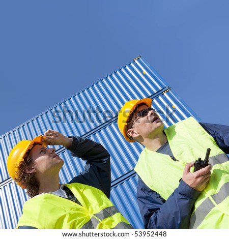 Two dockers, wearing a hard hat and safety vests at work under a blue sky, with huge containers in the background.