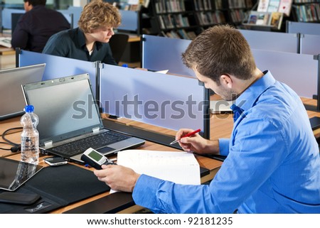 Student, writing down the answer from his calculator on his notepad, studying in a public library, with several other students in the background