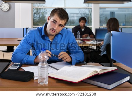 Student, looking into the camera, studying for his exams in a public library, with two other students in the background