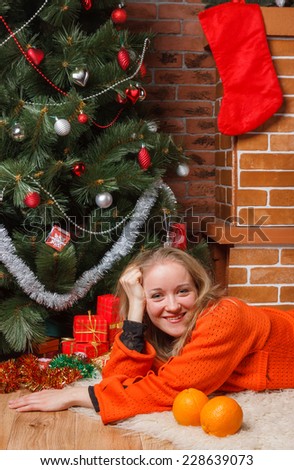 Smiling woman in orange sweater laying by decorated Christmas tree with oranges