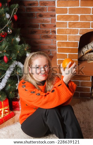 Beautiful blonde haired girl sitting next to decorated Christmas tree with orange in hands