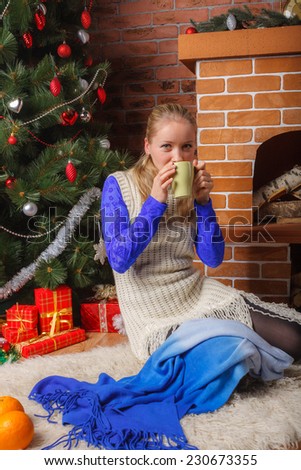 Beautiful woman in blue lace blouse and white knitted tunic drinking hot tea nearby Christmas tree
