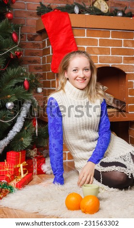 Blonde girl in a blue lace blouse and white sweater sitting by a Christmas tree with a cup of hot drink and a pair of oranges