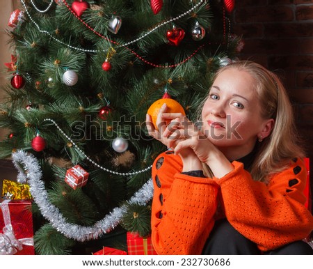 Beautiful woman sitting next to decorated Christmas tree with orange in hand