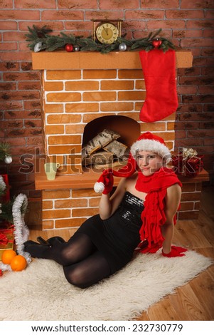 Young beautiful woman in red Santa hat and red scarf sitting by chimney on Christmas