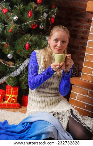 Young beautiful woman in blue lace blouse and white knitted tunic drinking hot coffee nearby Christmas tree