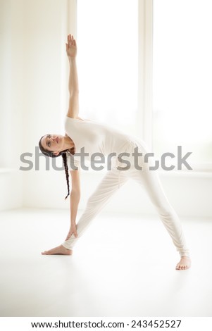 Young Caucasian woman doing yoga in sunny room. Trikonasana Triangle Pose.