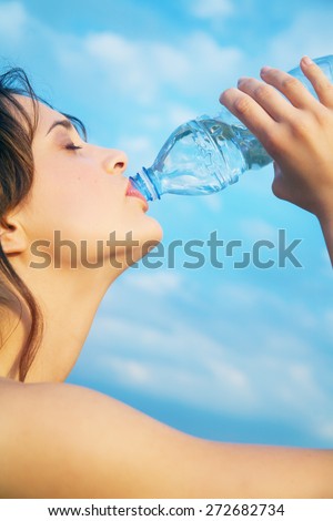 Young woman outdoor against blue sky drinking from bottle of mineral water.