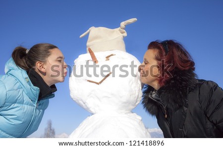 Cute mother and her daughter kissing snowman on a sunny winter day