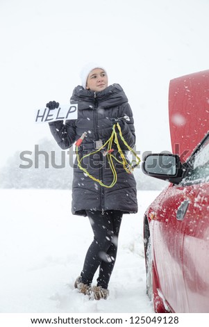 Cute girl waiting for help on the road holding HELP sign, she needs a battery boost for her car, on very cold, snowy winter day