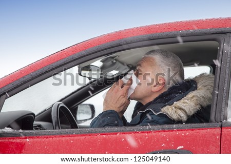 Mid aged man with flu symptoms sneezes in a car on snowy  winter day. Using handkerchief closes mouth with his hands