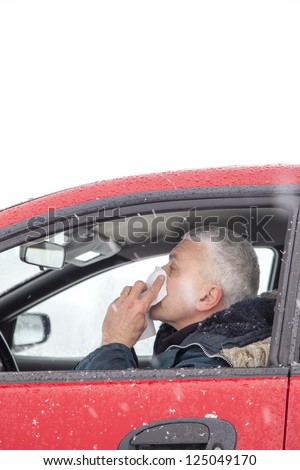 Mid aged man with flu symptoms sneezes in a car on snowy  winter day. Using handkerchief closes mouth with his hands