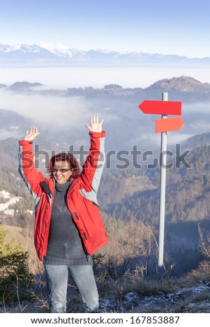 Cute smiling hiker woman at mountain top summit waving hello, standing by the red guidepost, space for text