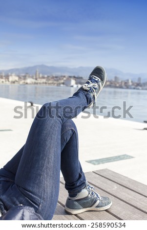 Laziness and relaxation. A pair of human legs in jeans pants and bright blue sport shoes with long shoelaces, on sea coast, against coastal town, selective focus on legs, space for text