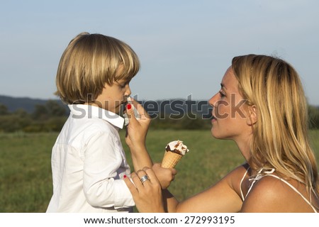 Young cute blonde boy eating a tasty ice cream, with his mother outdoors on sunny summer afternoon